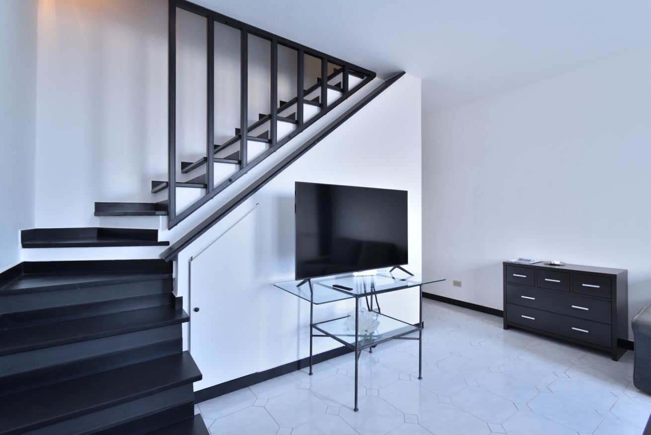 Un interior moderno con una escalera, un soporte de vidrio para televisor y un mueble de madera oscura.