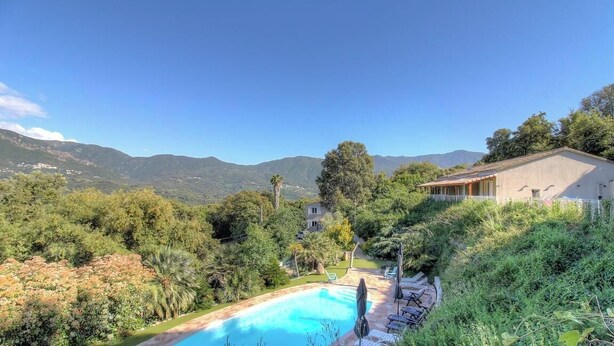 Una piscina rodeada de vegetación exuberante, una casa con techo inclinado y montañas al fondo.