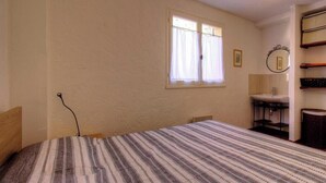 Une chambre à coucher avec un lit, une fenêtre avec des rideaux, une table de chevet, un miroir et une petite salle de bain avec un lavabo.
