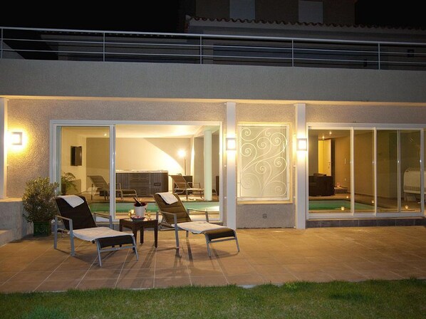 An outdoor patio with two lounge chairs, a small table, and a pool area visible through glass doors. - Santa Giulia - SOLENZARA (SOLENZARA)