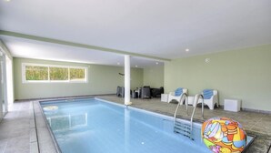 En indendørs swimmingpool med udsigt til grøn vegetation, et loungeområde med stole, og en farverig oppustelig bold.