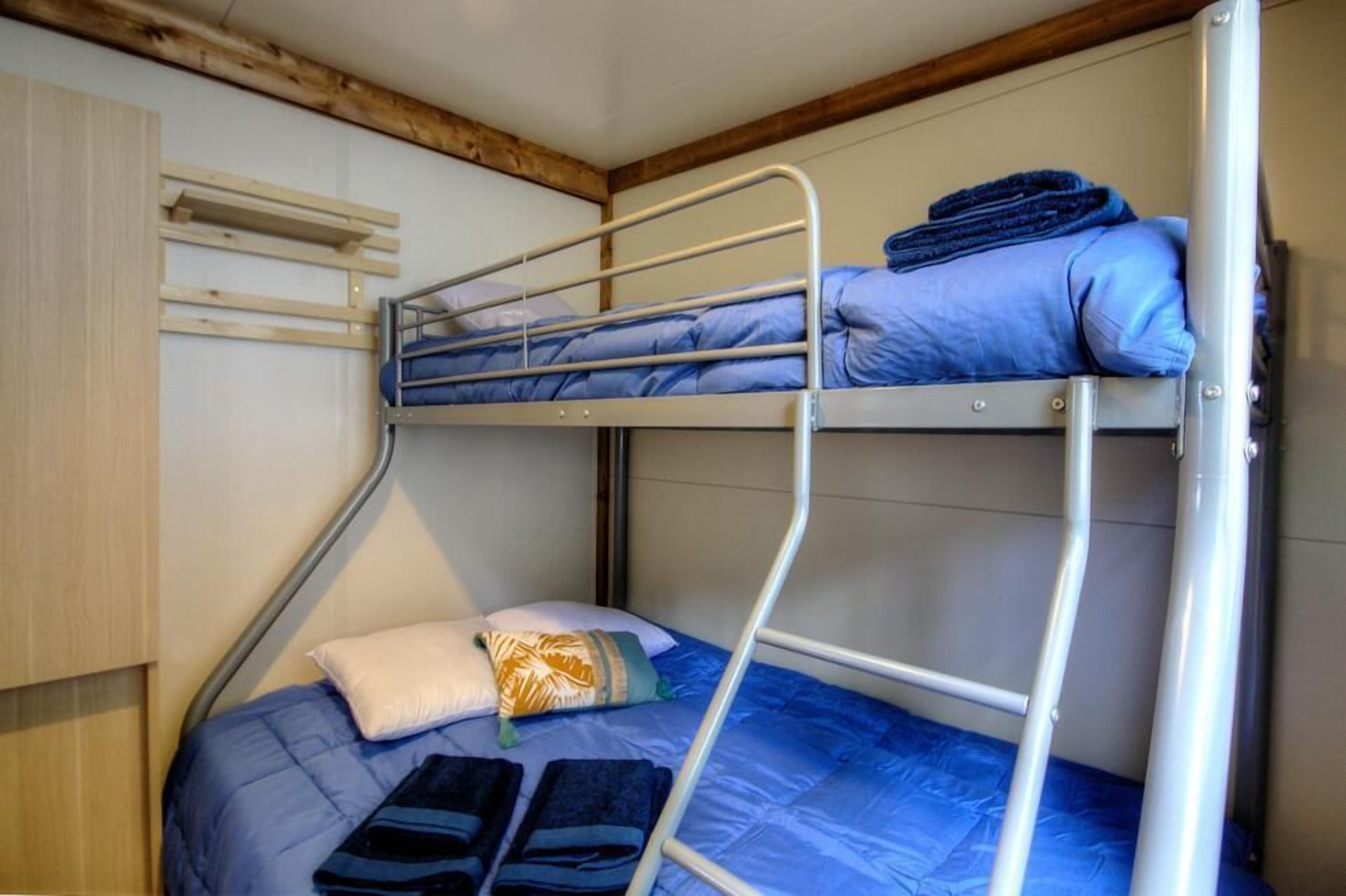 Een stapelbed met een blauwe matras, een houten hoofdbord en een metalen ladder.