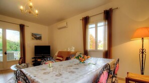Ein Zimmer mit Esstisch, Stühlen, Fernseher, Lampe und einem Fenster mit Vorhängen.