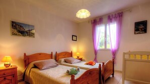 Un dormitorio con dos camas de madera, una cuna, una mesita de noche con lámpara y una ventana con cortinas moradas.