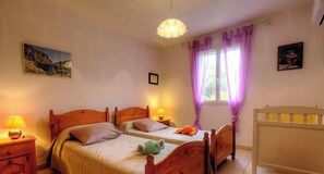 Ein Schlafzimmer mit zwei Holzbetten, einem Babybett, einem Nachttisch mit Lampe und einem Fenster mit violetten Vorhängen.