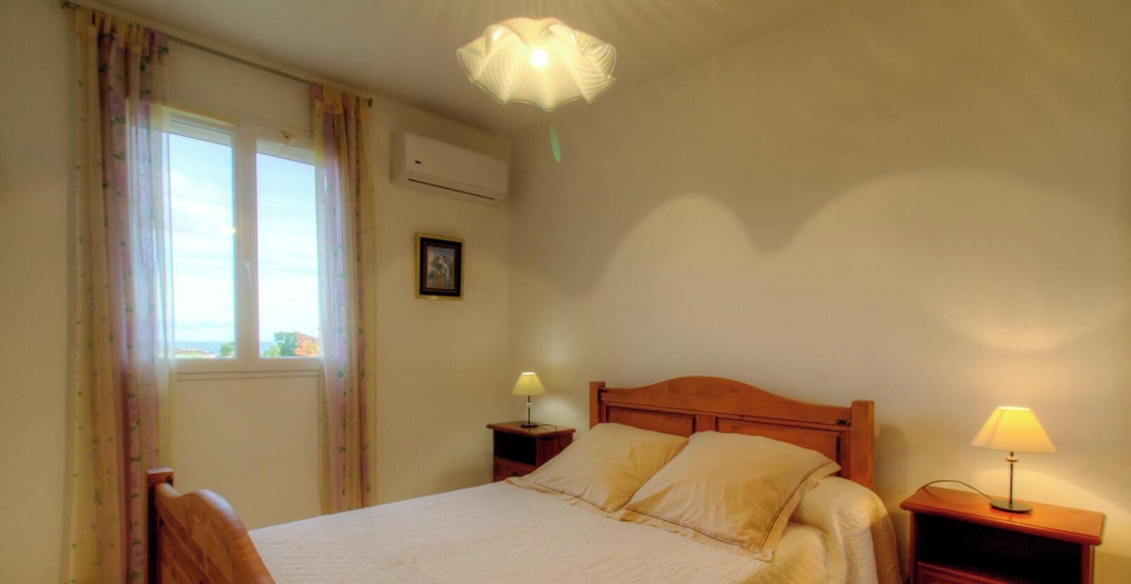 Ein Schlafzimmer mit einem Bett, zwei Nachttischen mit Lampen, einem Fenster mit Vorhängen, einer Klimaanlage und einem Wandbild.