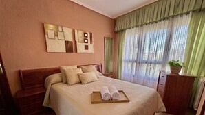 Una camera d'albergo con un letto, un comodino in legno, una cassettiera, una pianta in vaso e un'opera d'arte appesa al muro.