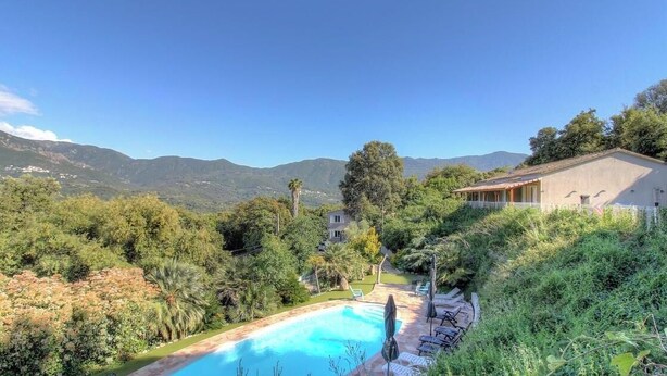 Una piscina rodeada de vegetación exuberante, una casa con techo inclinado y montañas al fondo.