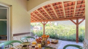 Una mesa de desayuno con pasteles, fruta y bebidas, bajo una pérgola de madera con vistas a zonas verdes y edificios.