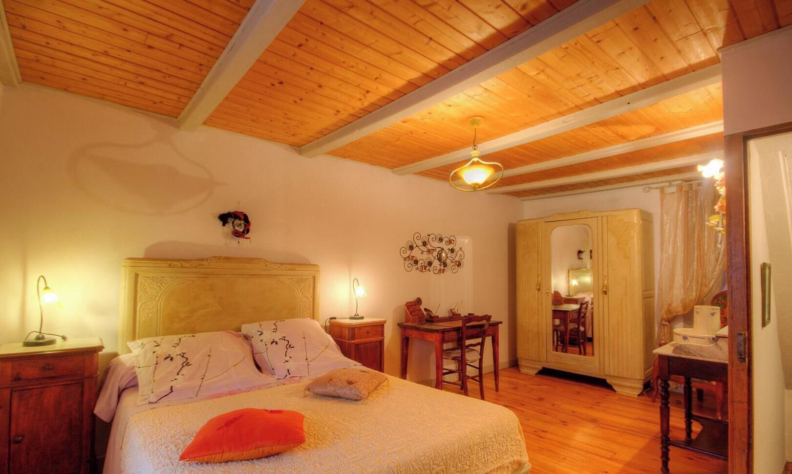 A bedroom with a wooden ceiling, a bed with pillows, a nightstand, a desk, and a wardrobe.