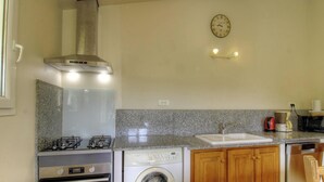 A compact kitchen with a gas stove, a built-in oven, a washing machine, wooden cabinets, a granite countertop, a stainless steel range hood, a wall clock, and a coffee maker. - Ogliastrello sheepfolds (FIGARI)