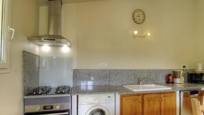 Eine kompakte Küche mit Gaskochfeld, Backofen, Waschmaschine, Holzschränken, einer Granit-Arbeitsplatte, einer Edelstahl-Dunstabzugshaube, einer Wanduhr und einer Kaffeemaschine.