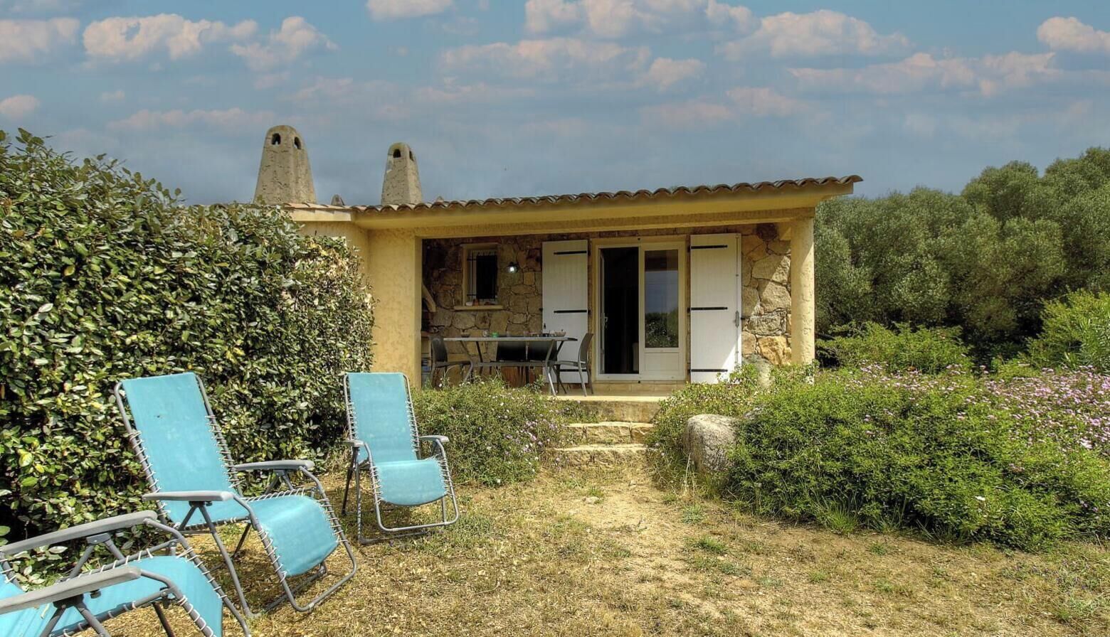 Een klein gebouw met een stenen gevel, een tafel en stoelen buiten en een tuin met struiken en een haag.