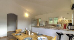 Ein Essbereich mit Holztisch, gedeckt für vier Personen, eine Küche mit Mikrowelle und Toaster sowie ein Fenster mit Blick auf Grünflächen.