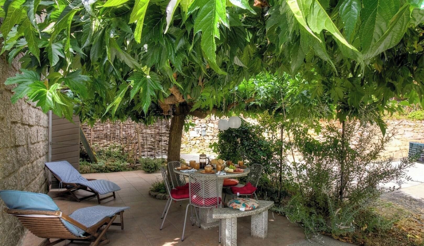 Un patio al aire libre con mesa y sillas, rodeado de follaje y un muro de piedra.