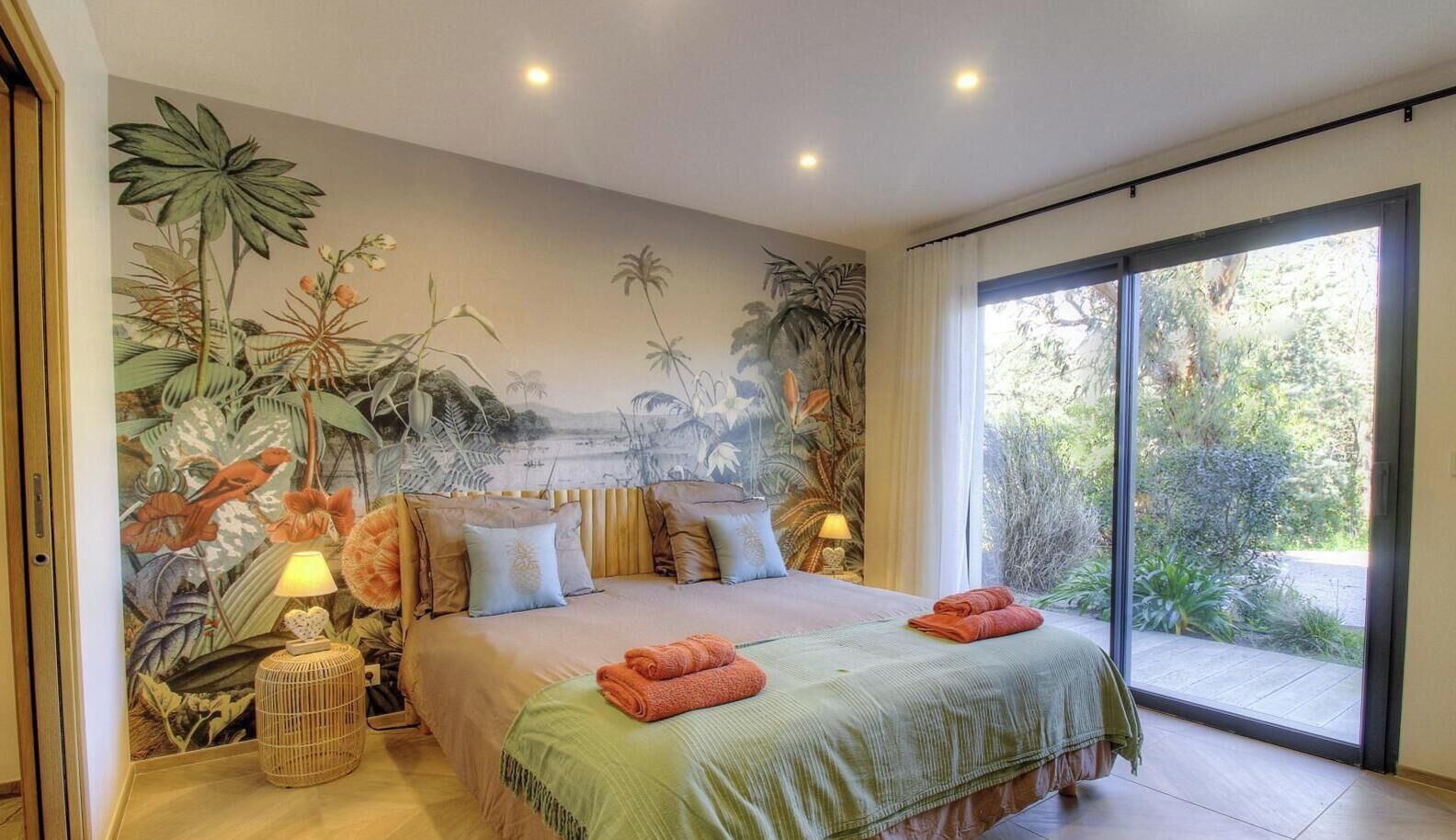 Una camera da letto con un letto grande, un affresco con piante tropicali, una porta scorrevole in vetro verso un giardino e un piccolo comodino con una lampada.