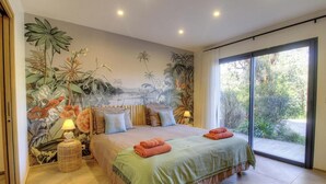 Ein Schlafzimmer mit einem großen Bett, einer Wandmalerei von tropischen Pflanzen, einer Schiebetür zu einem Garten und einem kleinen Nachttisch mit einer Lampe.