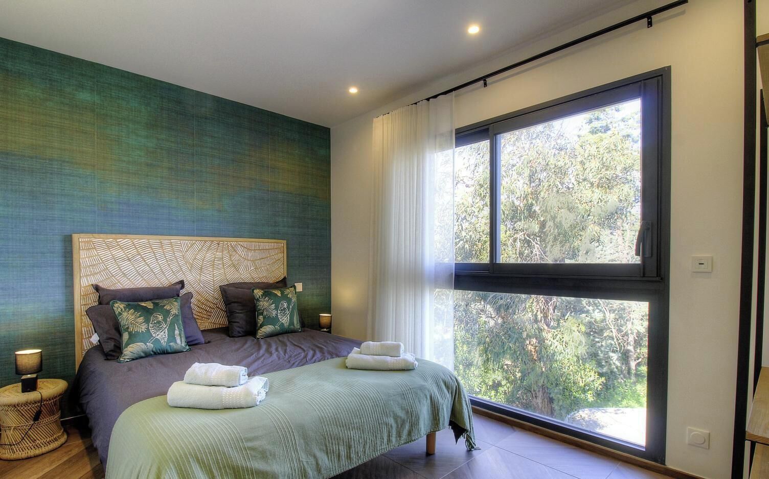 A bedroom with a large window, a bed with pillows, and a bedside table.