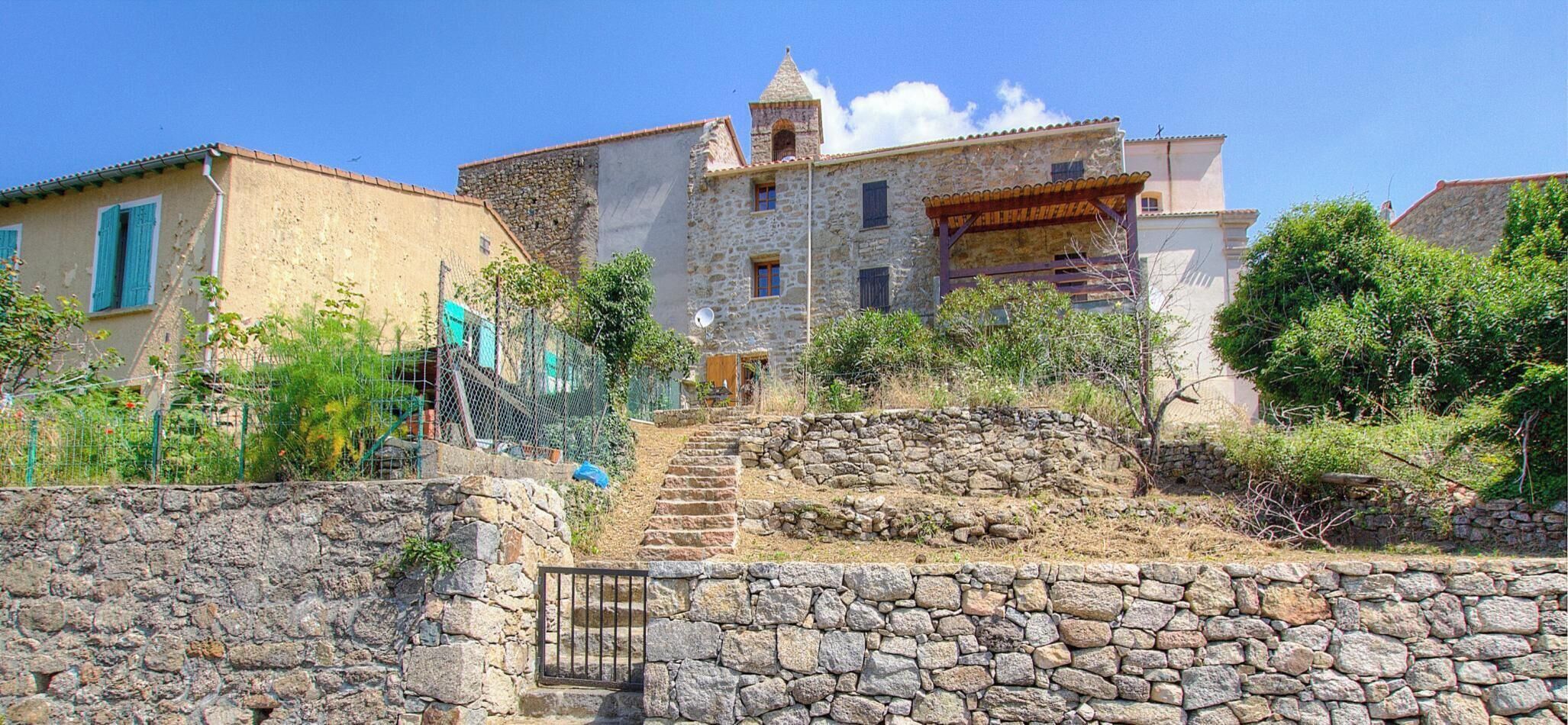 Una escalera de piedra que lleva a un edificio de piedra con una torre, rodeado de vegetación y un muro de piedra.