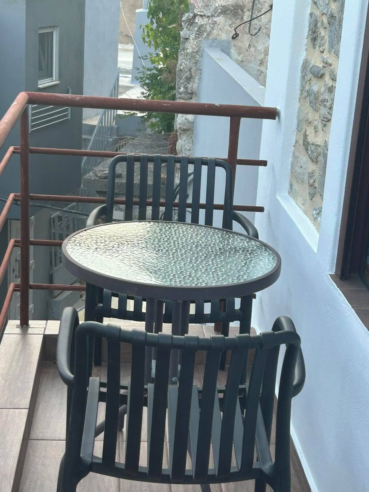 Un balcon avec une table ronde et des chaises, une rambarde en métal et une vue sur les bâtiments et la végétation.