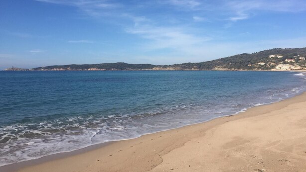 Ein Strand mit klarem, blauem Wasser, einem Sandufer und einer entfernten Hügelkette mit Gebäuden.