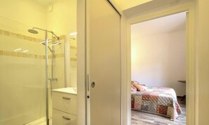 Un baño con cabina de ducha de vidrio, un lavabo blanco y una puerta que da a un dormitorio con cama y mesita de noche.
