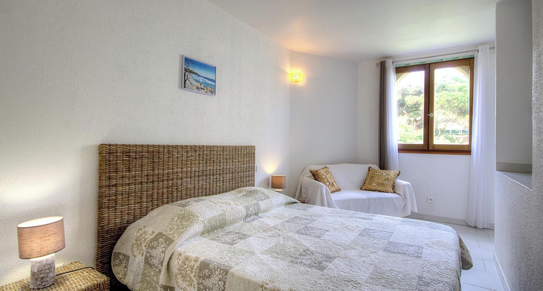 A bedroom with a bed, a bedside table, a lamp, a chair, a window, and a wall-mounted light fixture.