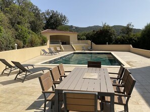 A pool with lounge chairs and a table, surrounded by a patio area with a view of hills. - The wild olive villa (SARTENE)