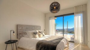 A bedroom with a large bed, a headboard with a geometric pattern, a hanging light fixture, and a view of a pool and mountains through large windows. - Villa Rocher - SAINTE LUCIE DE PORTO VECCHIO (SAINTE LUCIE DE PORTO VECCHIO)