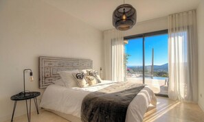 A bedroom with a large bed, a headboard with a geometric pattern, a hanging light fixture, and a view of a pool and mountains through large windows. - Villa Rocher - SAINTE LUCIE DE PORTO VECCHIO (SAINTE LUCIE DE PORTO VECCHIO)