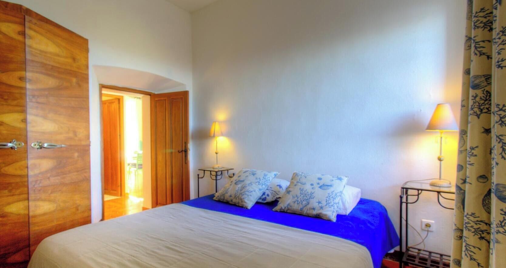 Quarto com cama, guarda-roupa de madeira, duas luminárias de cabeceira e uma porta que dá para outro cômodo.