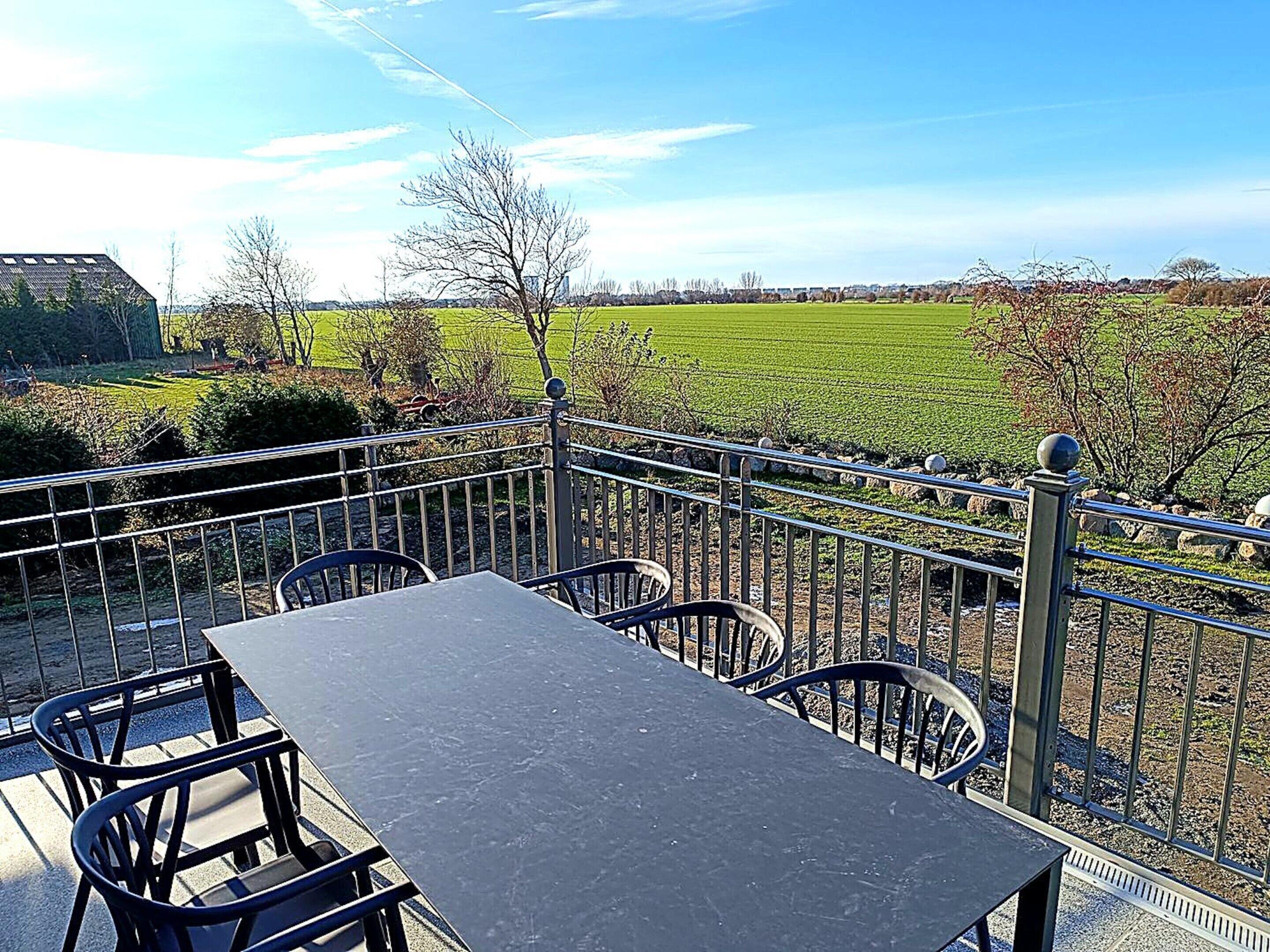 An outdoor patio with a table and chairs, a metal railing, and a view of a green field.
