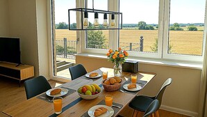 Ein Essbereich mit einem für ein Frühstück gedeckten Tisch, einem Fernseher und Blick auf ein Feld durch große Fenster.