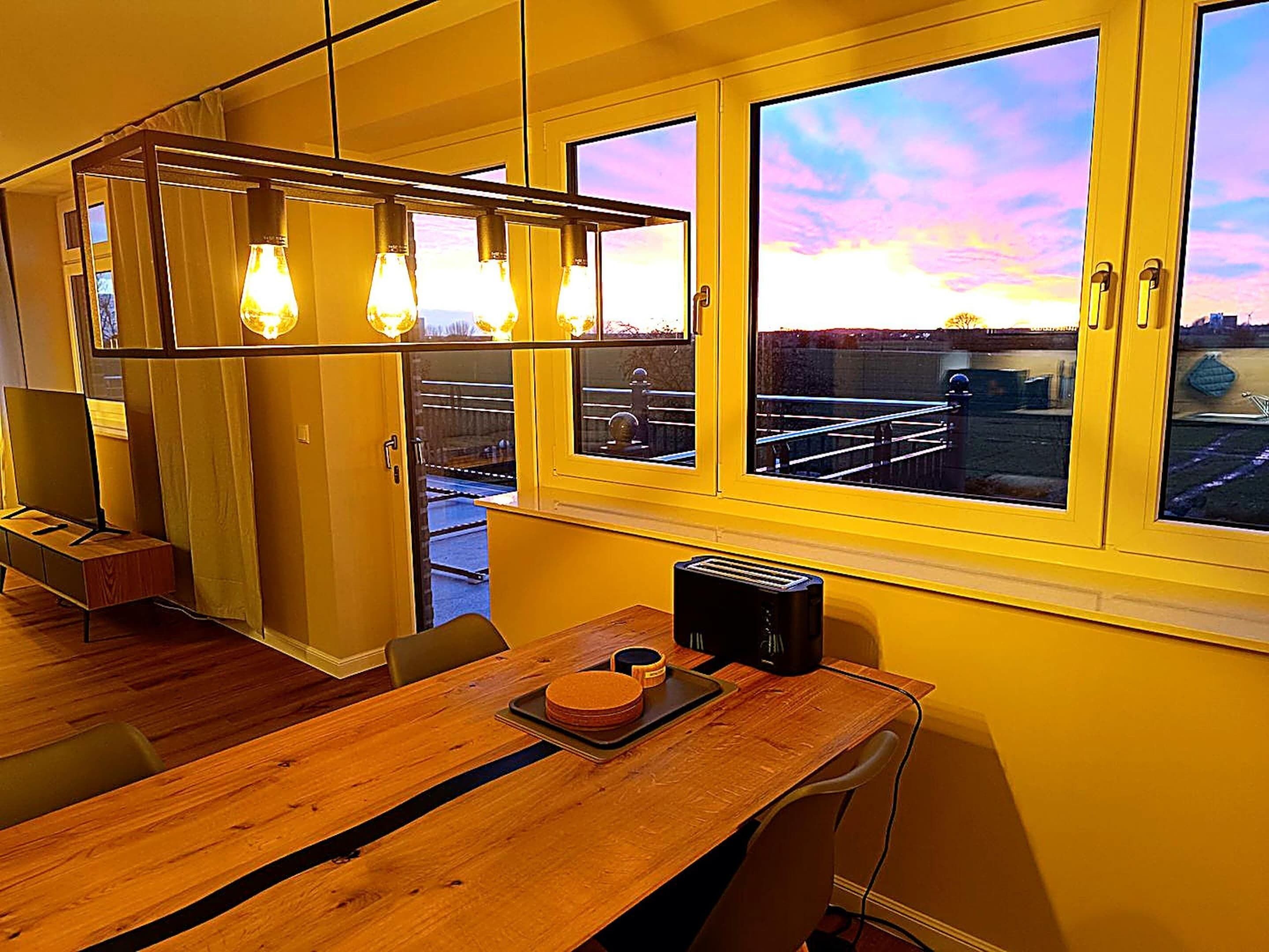 Ein Zimmer mit Holztisch, einem Fenster mit Blick auf eine Stadtlandschaft und einer Pendelleuchte.