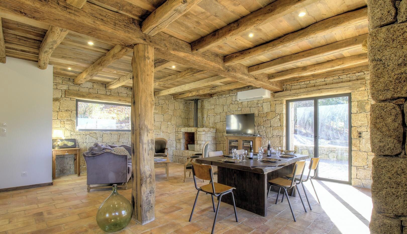 Un intérieur rustique avec des poutres apparentes, des murs en pierre, une cheminée en pierre, un coin repas avec des chaises et une grande fenêtre.