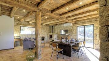 Un intérieur rustique avec des poutres apparentes, des murs en pierre, une cheminée en pierre, un coin repas avec des chaises et une grande fenêtre.