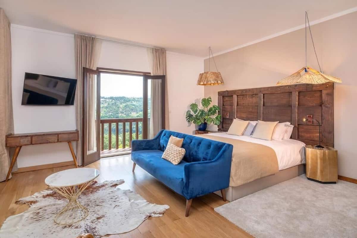 A bedroom with a large bed, a blue sofa, a small round table, a potted plant, and a view of the outdoors.