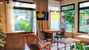 Een kamer met een houten plafond, een eettafel met stoelen, een schommelstoel en grote ramen met uitzicht op groen.