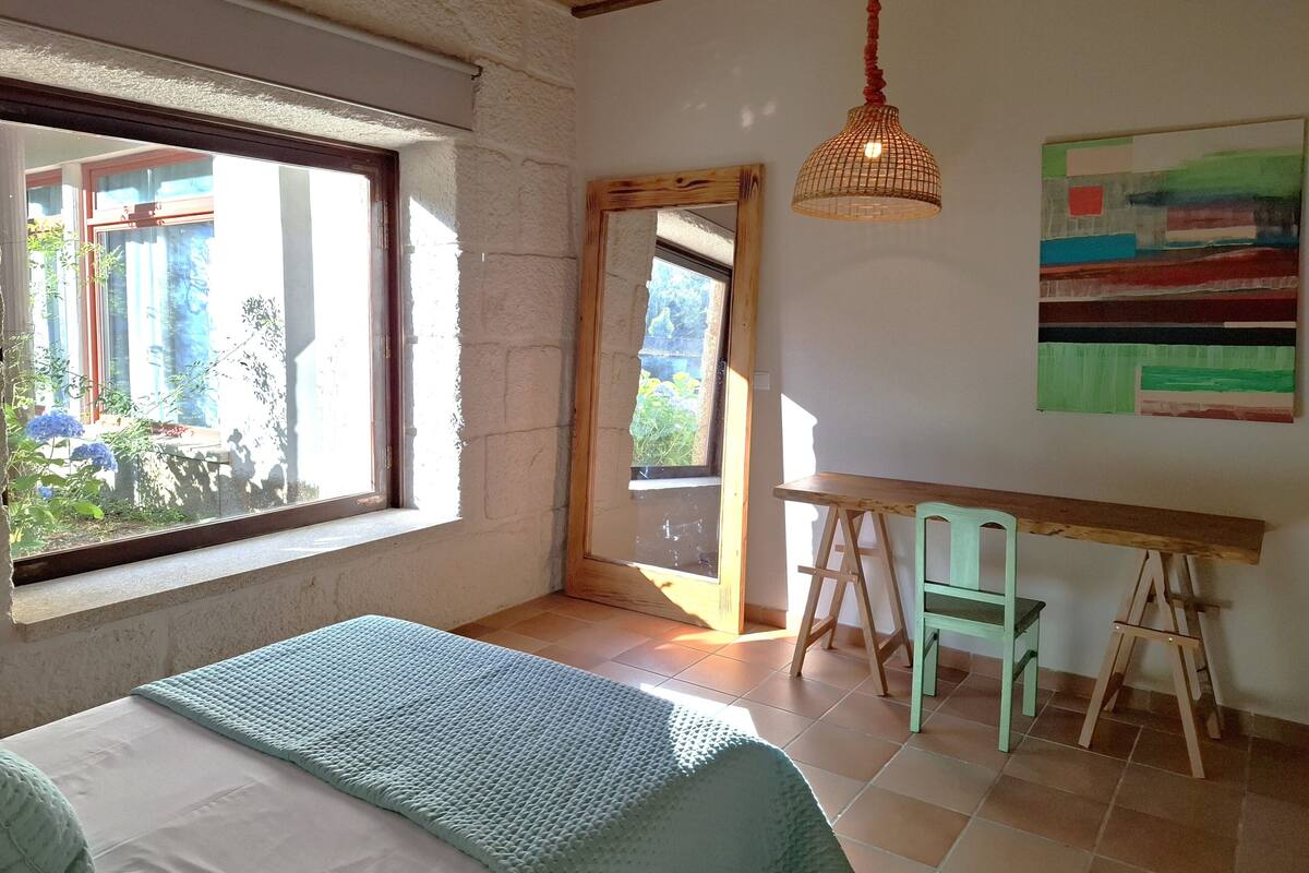A room with a bed, a desk, a chair, a hanging light fixture, and a painting on the wall.