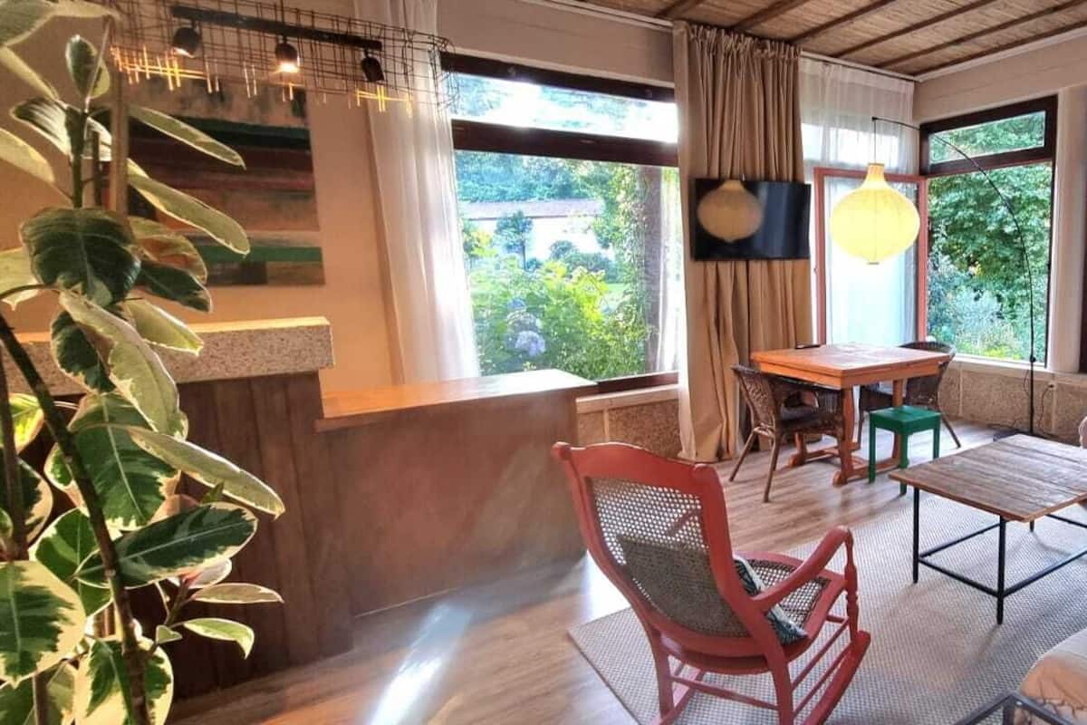 A room with a wooden desk, a red chair, a sofa, a table, and large windows with a view of greenery.