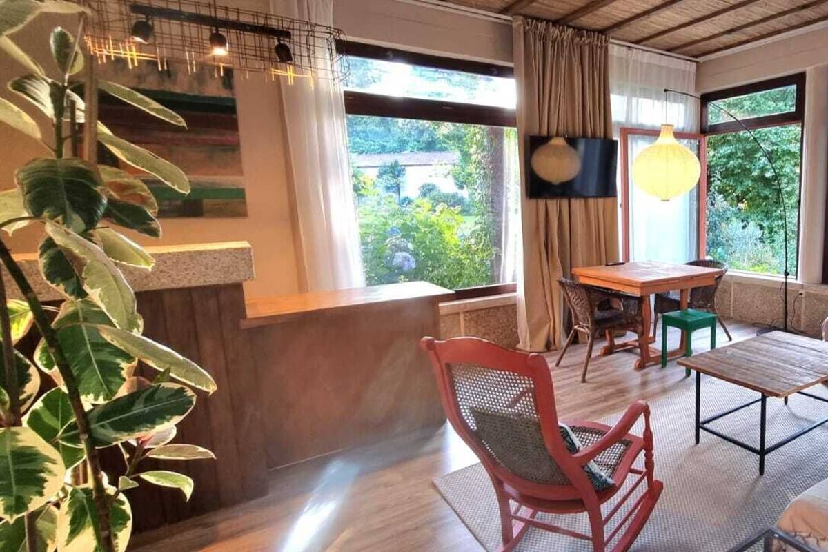 A room with a wooden desk, a red chair, a sofa, a table, and large windows with a view of greenery.