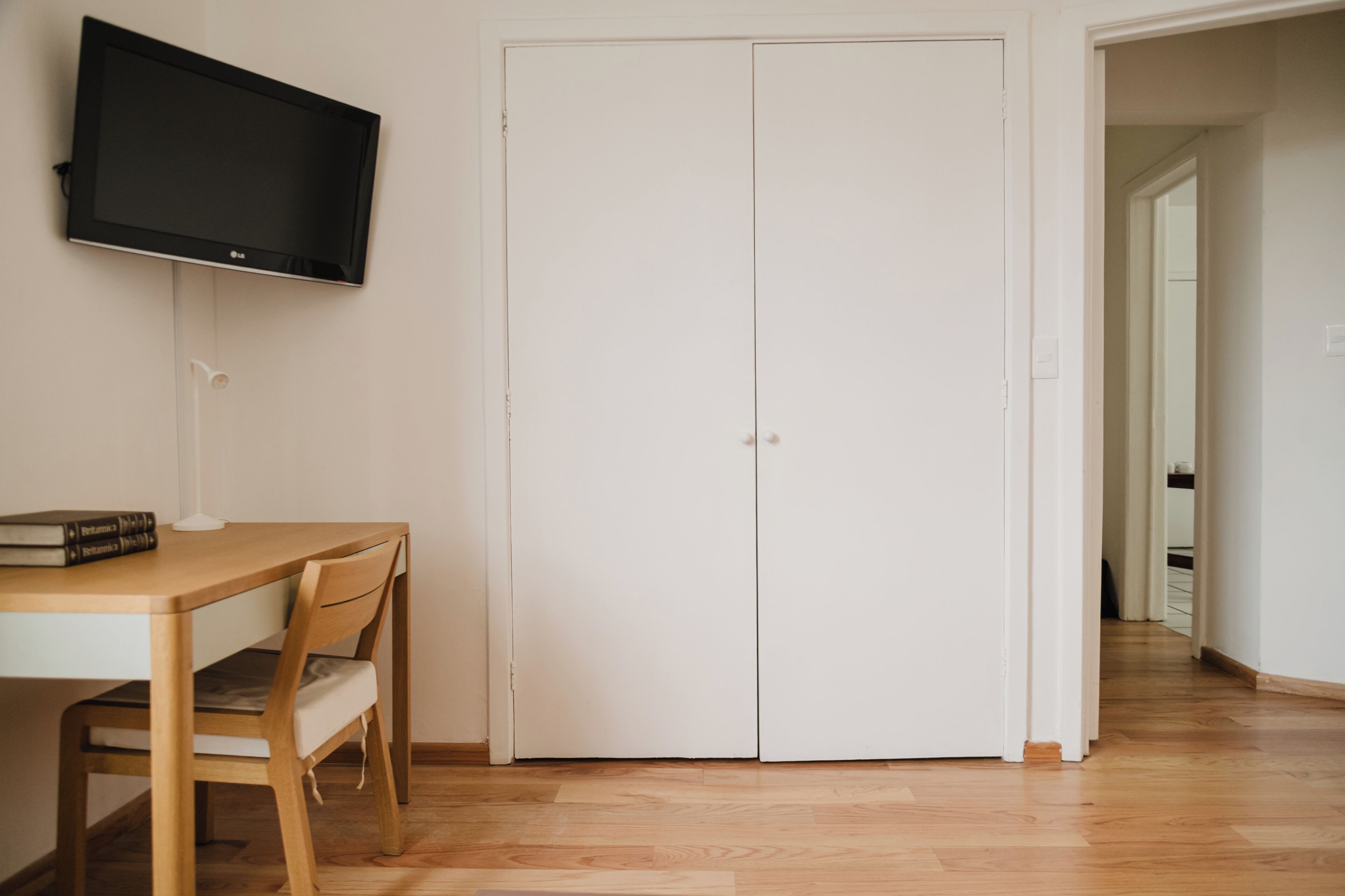 A room with a wooden desk, a television mounted on the wall, and a white door.