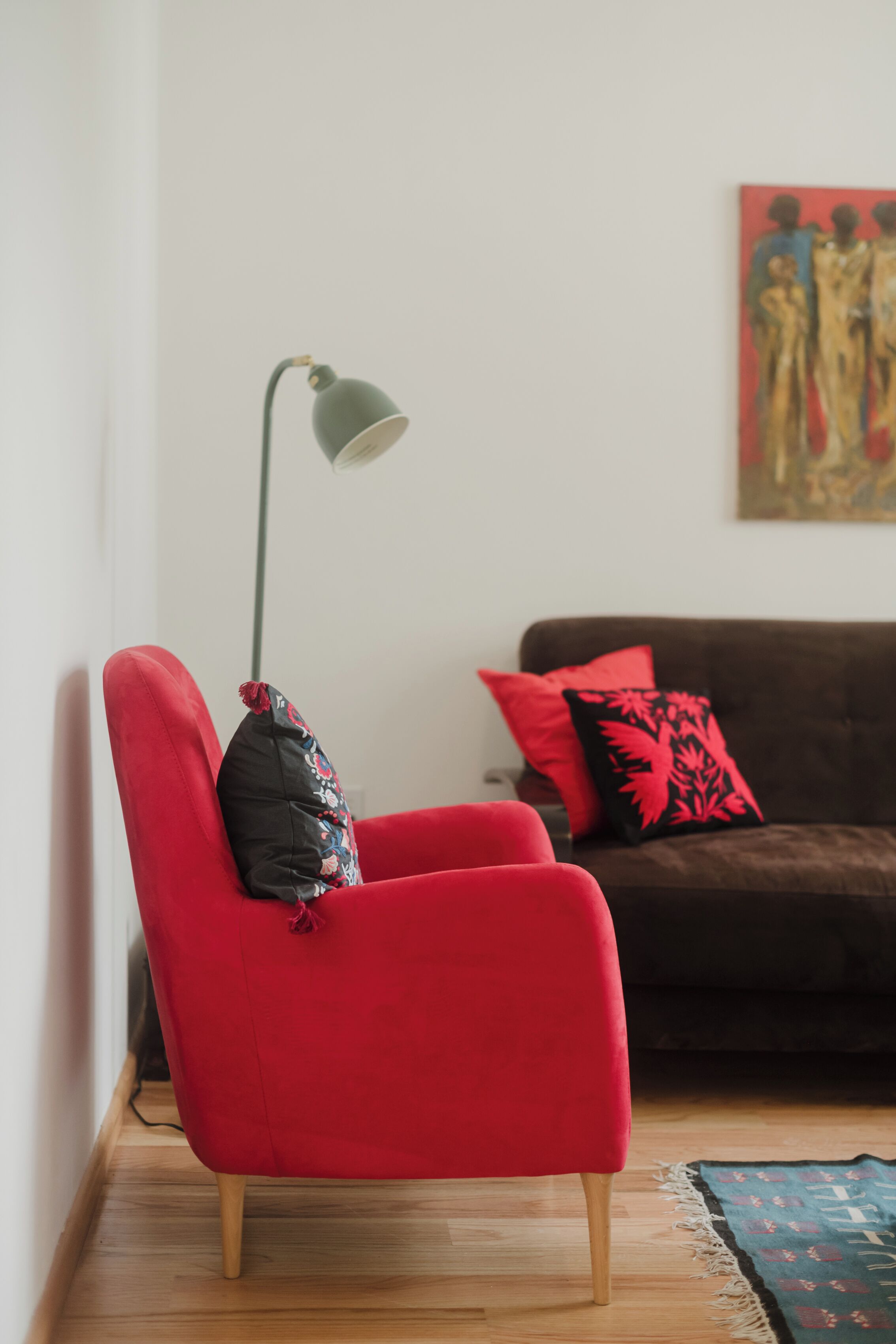 Un sillón rojo con un cojín negro, una lámpara de pie, un sofá marrón con cojines rojo y negro, y un cuadro en la pared.