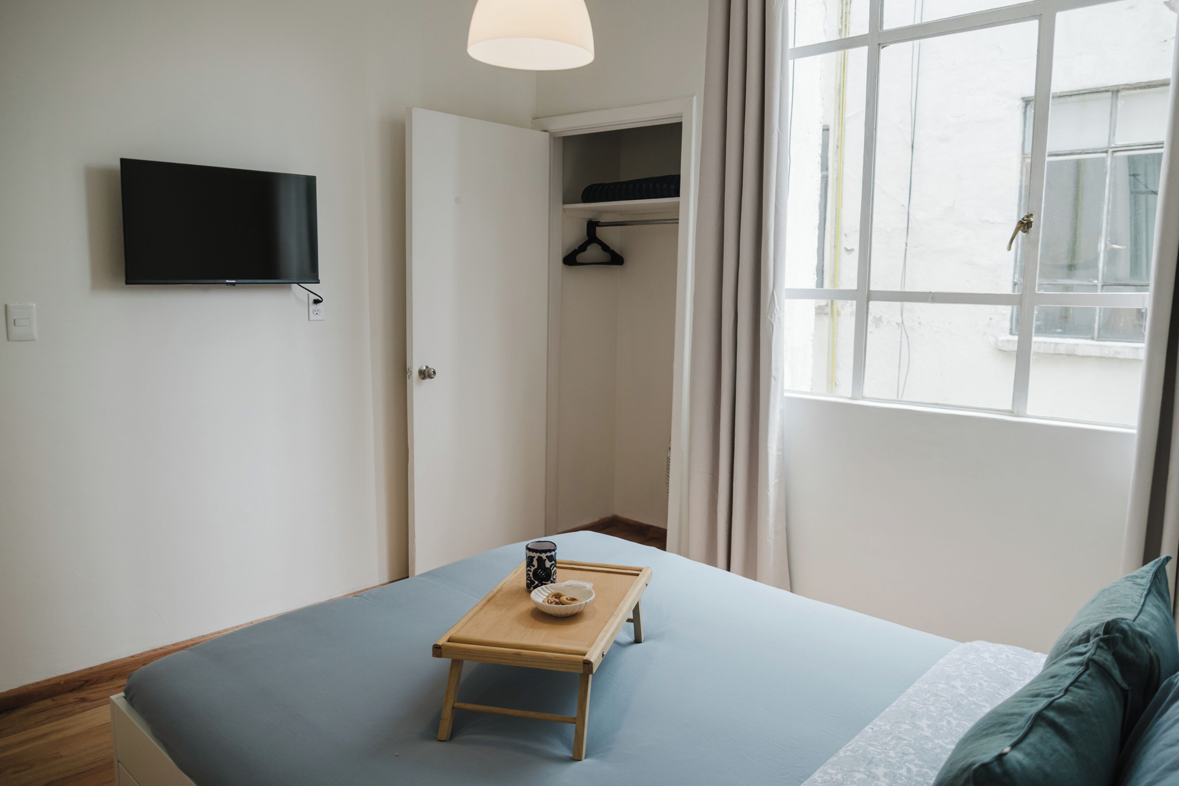 Une chambre avec un lit, un téléviseur fixé au mur, une petite table en bois avec un bol et une tasse, et une fenêtre avec des rideaux.