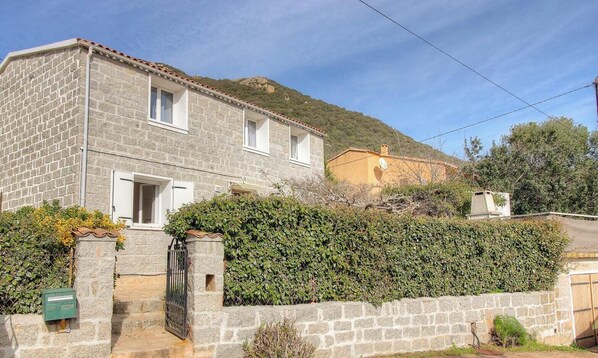Ein Haus aus Stein mit einem Ziegeldach, ein kleiner Garten und ein Hügel im Hintergrund.