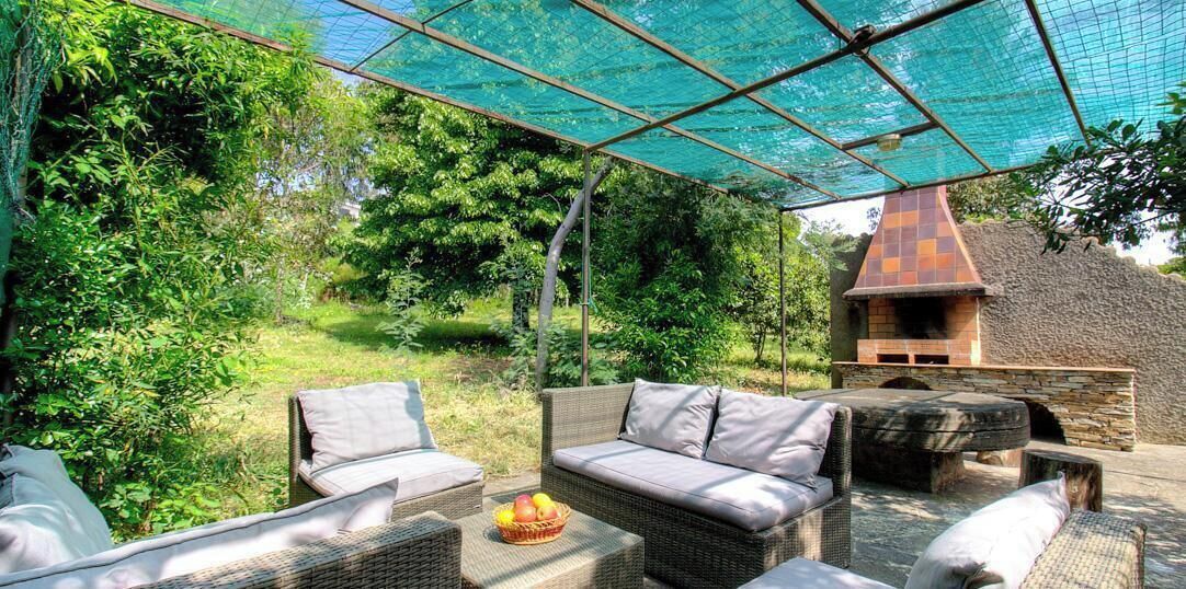 Une terrasse extérieure avec des meubles en osier, un barbecue en pierre et un filet vert tendu au-dessus.