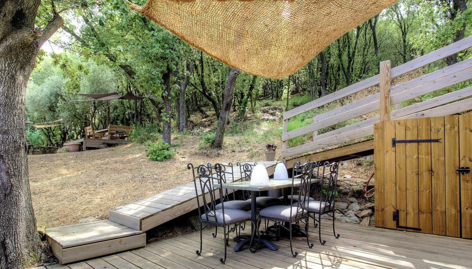 Un terrazzo in legno con tavolo e sedie, circondato da alberi e un recinto di legno.