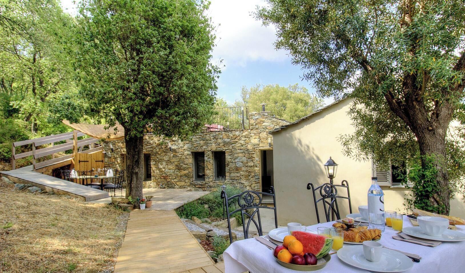 A rustic outdoor dining area with a stone building, wooden deck, and a table set for breakfast.