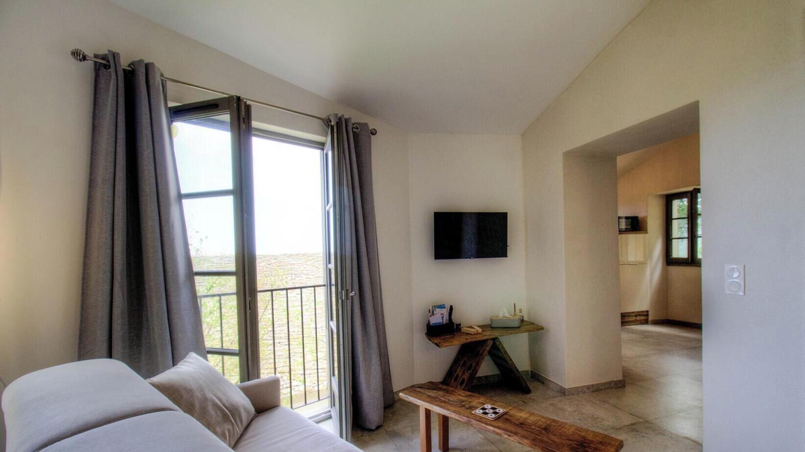 Ein Zimmer mit einer Couch, einem kleinen Tisch, einem Fernseher und einem Balkon mit Vorhängen.