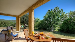 Eine überdachte Terrasse mit Frühstückstisch, umgeben von Stühlen und Blick auf einen üppigen Garten.